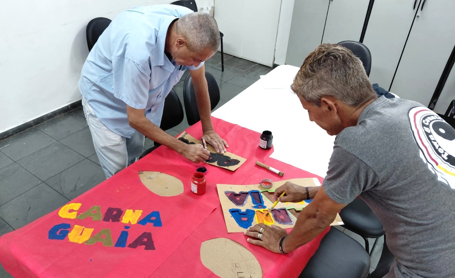 A imagem mostra dois homens trabalhando juntos na confecção de um cartaz artesanal, sobre uma mesa coberta por papel vermelho. Eles estão pintando letras e formas coloridas com pincéis e tinta, usando moldes recortados em papelão.  No cartaz, é possível ler a palavra “Carna Guaia”, escrita em letras grandes e coloridas. Sobre a mesa, há potes de tinta, pincéis e materiais de apoio para a pintura.  O ambiente parece ser uma sala simples, com cadeiras ao redor e paredes claras, indicando um espaço de oficina, reunião ou atividade comunitária. A cena transmite colaboração, criatividade e envolvimento em uma atividade coletiva