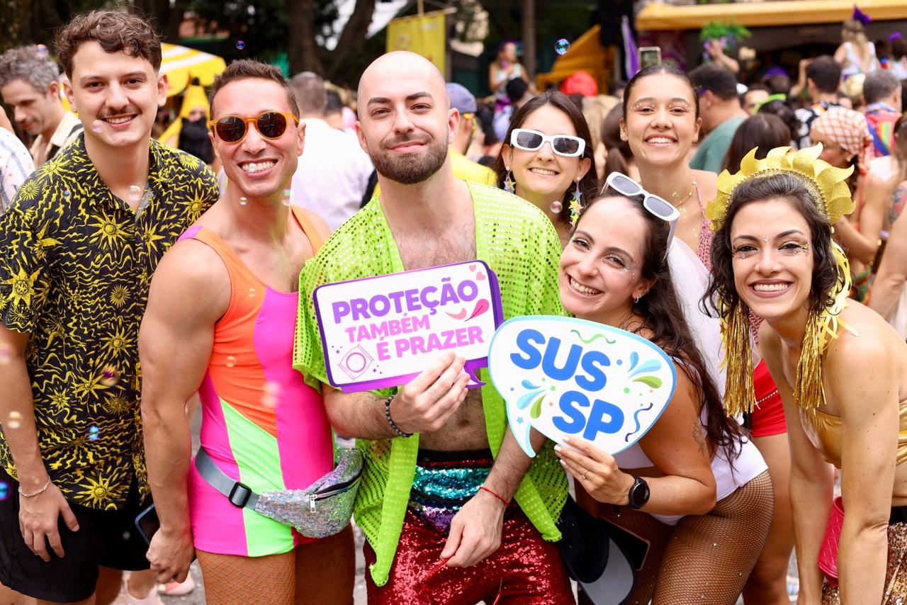 A imagem mostra um grupo de oito pessoas posando juntas em meio a um evento festivo ao ar livre, provavelmente um bloco de Carnaval.  Todos estão sorrindo e olhando para a câmera, demonstrando entusiasmo e alegria. As roupas são chamativas, coloridas e cheias de brilho, com destaque para peças em neon, paetês, transparências e acessórios divertidos, típicos de festas carnavalescas.  No centro, um homem segura uma placa com a frase “Proteção também é prazer”, acompanhada de um desenho de preservativo, reforçando uma mensagem de conscientização sobre saúde e prevenção. Ao lado dele, uma mulher segura outra placa com os dizeres “SUS SP”, indicando referência ao sistema público de saúde.
