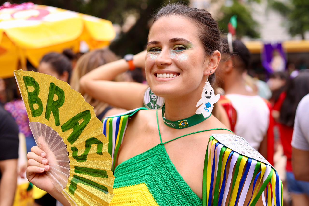 A imagem mostra uma mulher sorridente em primeiro plano, participando de um evento festivo ao ar livre, provavelmente um bloco de Carnaval.  Ela olha diretamente para a câmera e demonstra alegria. Usa maquiagem colorida nos olhos, com tons de verde e brilho no rosto, além de glitter abaixo dos olhos. O cabelo está preso, deixando o rosto em evidência.  Ela veste uma roupa nas cores verde, amarelo e azul, remetendo à bandeira do Brasil, com franjas coloridas nos ombros. No pescoço, usa um acessório verde com a palavra “Brasil” e um pequeno símbolo da bandeira. Também chama atenção um brinco em formato de fantasma e outro com estampa.