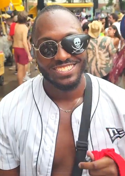 A imagem mostra um homem sorrindo em meio a um ambiente festivo ao ar livre, provavelmente durante um evento como um bloco de Carnaval.  Ele está em primeiro plano, olhando diretamente para a câmera, com uma expressão alegre e descontraída. Usa óculos escuros diferentes em cada lente: uma transparente e outra com o desenho de uma caveira, o que dá um toque divertido e estilizado ao visual.  Ele veste uma camisa branca com listras verticais, parcialmente aberta, deixando o peito à mostra, e carrega uma bolsa transversal no ombro. Também segura um pano ou peça de roupa vermelha na mão.
