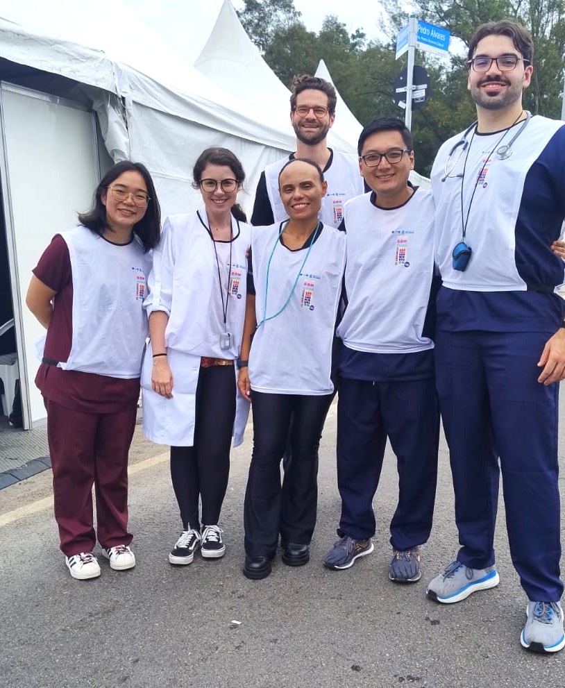 A imagem mostra um grupo de sete pessoas posando juntas em um ambiente externo, possivelmente em uma área de atendimento ou apoio durante um evento.  Todas vestem camisetas brancas com logotipo institucional, sugerindo que fazem parte de uma equipe organizada, provavelmente da área da saúde. Alguns usam estetoscópios, crachás ou colares de identificação, reforçando essa impressão.  O grupo está em frente a uma grande tenda branca, que pode ser um posto de atendimento temporário. Ao fundo, há árvores e placas de sinalização, indicando que o local é um espaço público, como um parque ou avenida.