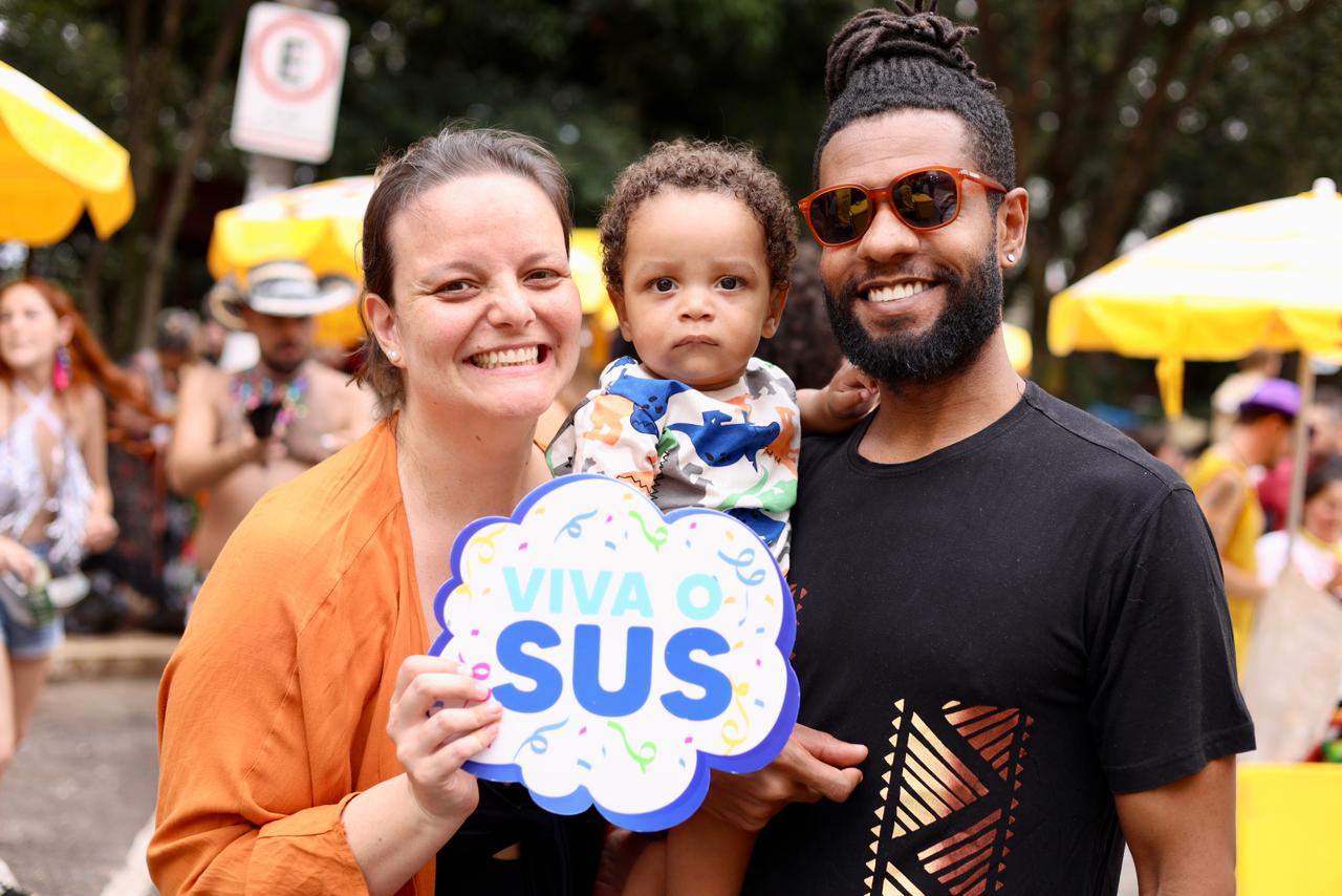 A imagem mostra três pessoas em primeiro plano, em meio a um ambiente festivo ao ar livre, provavelmente durante um bloco de Carnaval.  À esquerda, há uma mulher sorridente, usando uma blusa laranja, que segura uma placa com a frase “Viva o SUS”, em letras azuis e com detalhes coloridos ao redor. Ela olha diretamente para a câmera, transmitindo alegria e simpatia.  No centro, há uma criança pequena sendo segurada pelos adultos. A criança veste uma roupa estampada com desenhos coloridos e tem uma expressão mais séria, contrastando com o clima alegre ao redor.  À direita, aparece um homem sorridente, usando óculos escuros de armação marrom, camiseta preta com estampa geométrica dourada e cabelo preso em dreadlocks. Ele também olha para a câmera, demonstrando descontração.