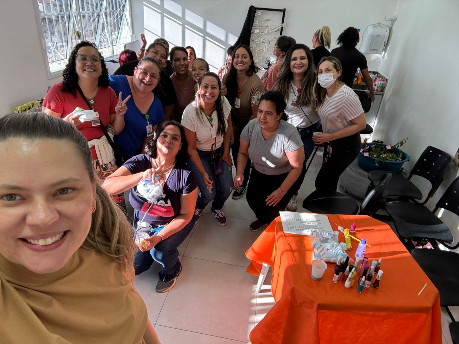 A imagem mostra um grupo de mulheres reunidas em um ambiente interno bem iluminado, provavelmente em uma sala de unidade de saúde ou espaço comunitário. A foto foi tirada em estilo selfie, com uma mulher em primeiro plano à esquerda, sorrindo para a câmera.  Ao fundo, cerca de 15 a 20 mulheres também sorriem e posam de forma descontraída, algumas fazendo sinal de “paz” com as mãos. Muitas usam crachás, o que sugere que sejam profissionais ou participantes de alguma atividade institucional. Algumas vestem máscaras, indicando cuidado com a saúde.  No centro e à direita da imagem, há uma mesa coberta com uma toalha laranja, sobre a qual estão dispostos diversos frascos coloridos, copos descartáveis e materiais que parecem ser utilizados em uma oficina ou atividade prática, possivelmente relacionada a autocuidado, beleza ou saúde.