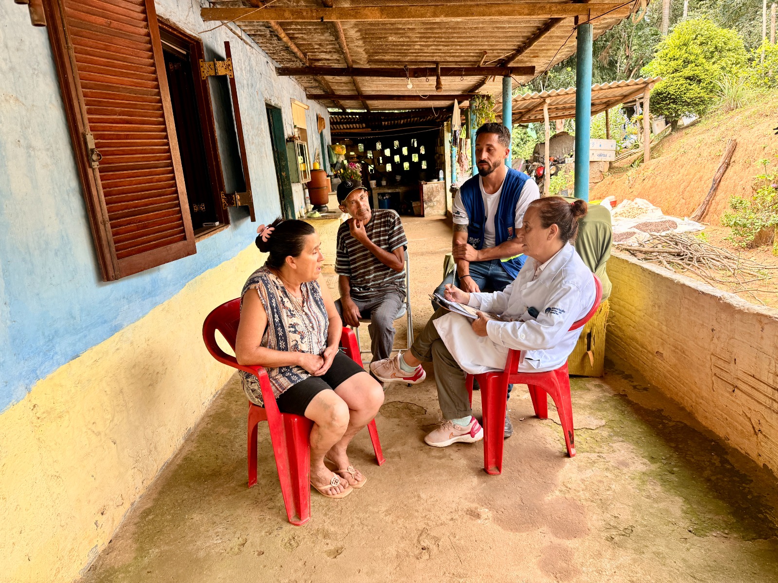 A imagem mostra uma visita domiciliar em uma área rural. Em uma varanda simples, com chão de cimento e cobertura de madeira, dois profissionais de saúde conversam com um casal de moradores.  Uma mulher está sentada em uma cadeira plástica vermelha, de frente para uma profissional que usa jaleco e anota informações em uma prancheta, indicando atendimento ou acompanhamento de saúde. Ao lado, um homem mais velho, também sentado, observa a conversa. Um agente comunitário de saúde, identificado pelo colete azul do SUS, está em pé próximo ao grupo, acompanhando o atendimento.  O ambiente é acolhedor e cotidiano, com elementos típicos de uma casa rural ao fundo, como utensílios domésticos e área externa com vegetação. A cena transmite proximidade, escuta e cuidado, característicos do trabalho da atenção básica e das equipes de saúde da família no território.