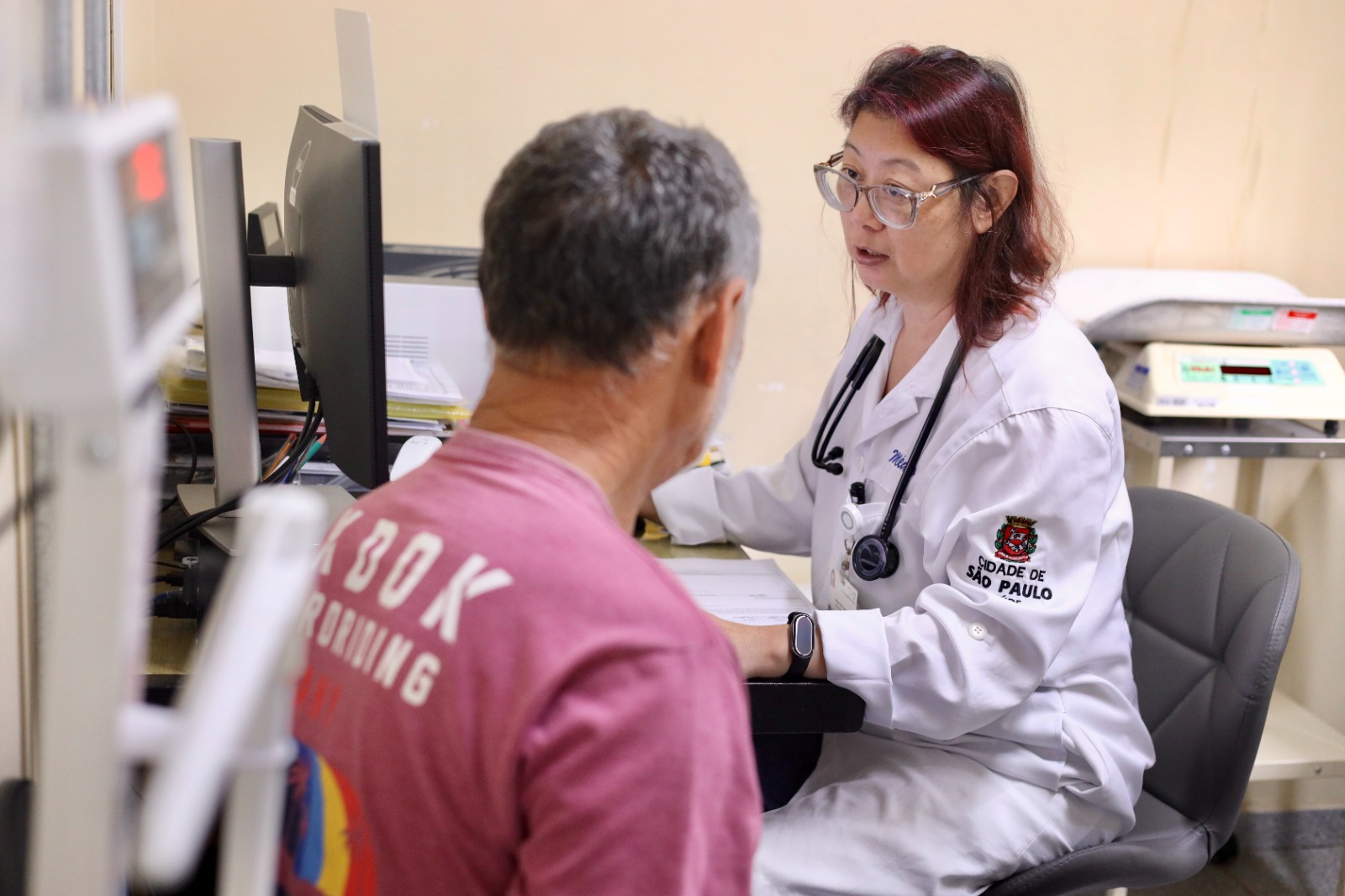 A imagem mostra uma consulta médica em um ambiente clínico. Uma profissional de saúde, usando jaleco branco com identificação da Prefeitura de São Paulo e um estetoscópio no pescoço, atende um paciente sentado à sua frente.  A médica está voltada para o paciente, falando e aparentemente explicando alguma informação, enquanto utiliza o computador e consulta documentos sobre a mesa. O paciente, um homem de meia-idade ou idoso, está de costas para a câmera e ouve atentamente.  O consultório é simples e funcional, com equipamentos médicos ao fundo e materiais organizados na mesa. A cena transmite um momento de escuta e orientação, típico do atendimento na atenção básica, com foco no acompanhamento e cuidado contínuo do paciente.