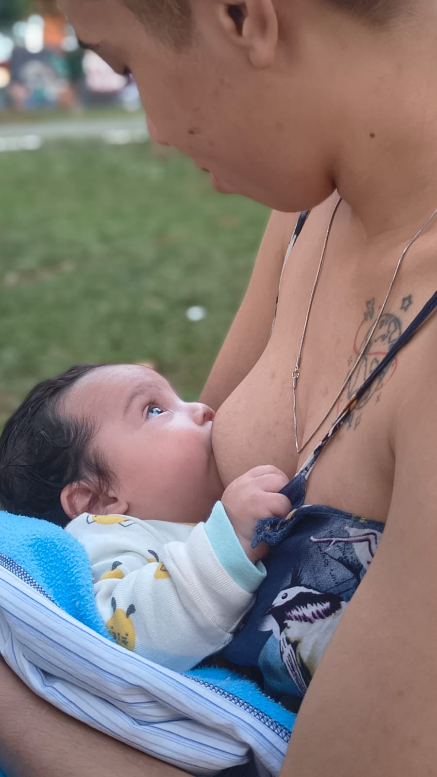 A imagem mostra uma mãe amamentando o bebê ao ar livre. O bebê está aconchegado no colo, mamando e olhando para a mãe, enquanto ela o segura junto ao corpo. A cena destaca o vínculo afetivo entre mãe e filho e a amamentação como um momento de cuidado, nutrição e proximidade.