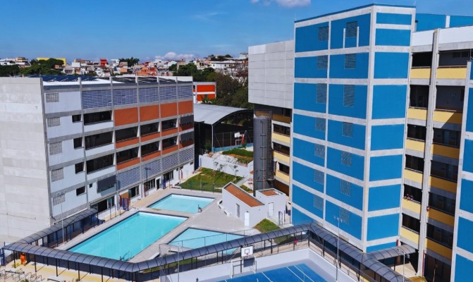 Vista de um conjunto educacional urbano com prédios modernos de vários andares. À direita, há um edifício alto com fachada em tons de azul e branco, com janelas organizadas em linhas. À esquerda, outro prédio apresenta faixas horizontais em cinza e laranja. Entre os edifícios, há áreas abertas com duas piscinas retangulares e uma quadra esportiva cercada. Ao fundo, observa-se o entorno urbano com casas e outros edifícios sob céu claro.