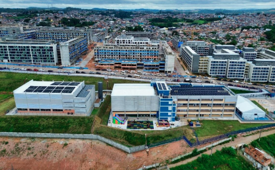 Vista aérea de um complexo urbano com edifícios institucionais e residenciais. Em primeiro plano, há um conjunto de prédios modernos, incluindo um edifício com painéis solares no telhado e outro com fachada azul e branca, cercados por área verde e cercamento. Ao fundo, observa-se uma ampla área urbana com diversos prédios, ruas movimentadas e bairros residenciais que se estendem até regiões de relevo elevado. O cenário indica um projeto urbano de grande escala integrado à cidade.