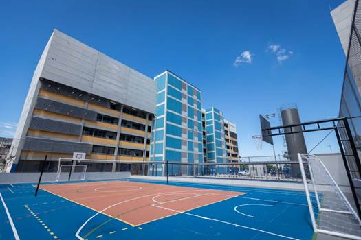 Imagem de um edifício de cinco andares, com quadra poliesportiva no primeiro plano.