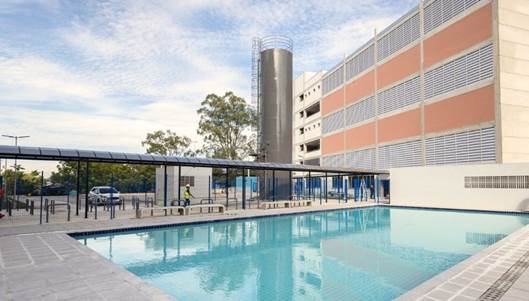 Imagem de um edifício de três andares, com piscina no primeiro plano e um corredor conectando ao edifício.