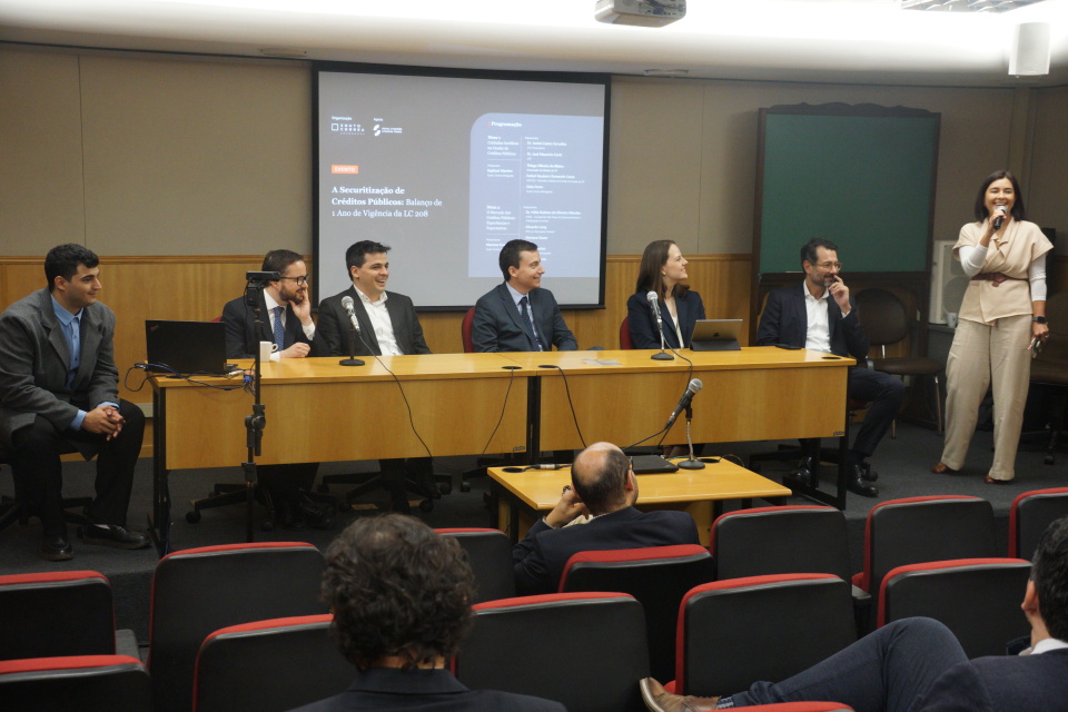 Registro do evento “A Securitização de Créditos Públicos: Balanço de 1 Ano de Vigência da LC 208”, realizado em auditório. A mediadora do debate está de pé à direita, segurando um microfone e falando ao público. À frente, seis participantes estão sentados atrás de uma mesa de madeira clara, prestando atenção à fala da mediadora, entre eles está o Diretor Presidente da SPDA. Ao fundo, uma tela projeta o título e a programação do evento. Na plateia, pessoas assistem atentamente à exposição. O ambiente é formal, com disposição típica de mesa-redonda institucional.