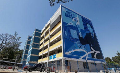 Imagem de um edifício de cinco andares, com um grafite na empena, de tons azulados, com a imagem do Silvio Santos, TV, rádio e aviões.