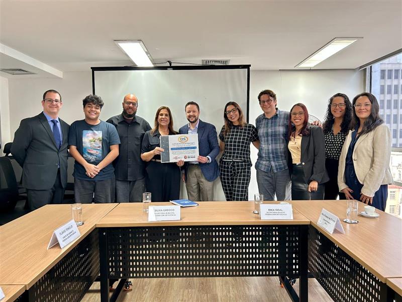 Dez pessoas posam em uma sala de reuniões, atrás de uma mesa em formato de “U”. Ao centro, duas seguram um certificado emoldurado e sorriem para a câmera. Sobre a mesa há copos com água e placas de identificação. Ao fundo, vê-se uma tela de projeção e janelas laterais que iluminam o ambiente.