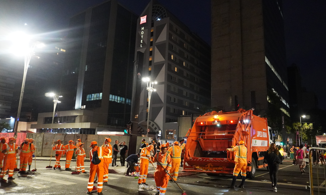 A fotografia noturna exibe uma cena urbana movimentada, com uma grande concentração de trabalhadores da limpeza pública vestidos com uniformes laranja vibrante e coletes refletivos. Eles estão espalhados por uma rua larga e escura, muitos segurando vassouras e outros equipamentos de limpeza.  Ao fundo, a avenida está iluminada por postes de luz e semáforos, alguns deles vermelhos. Carros com faróis e lanternas acesas formam uma fila no centro da imagem, sugerindo tráfego ou veículos parados. À direita, um prédio alto se destaca com sua fachada iluminada por luzes azuis verticais e horizontais, além de algumas janelas acesas. Mais ao fundo, outros edifícios da cidade também exibem luzes, criando um cenário de metrópole à noite. O ambiente é de atividade noturna intensa, com os garis realizando seu trabalho em meio ao cenário urbano iluminado.