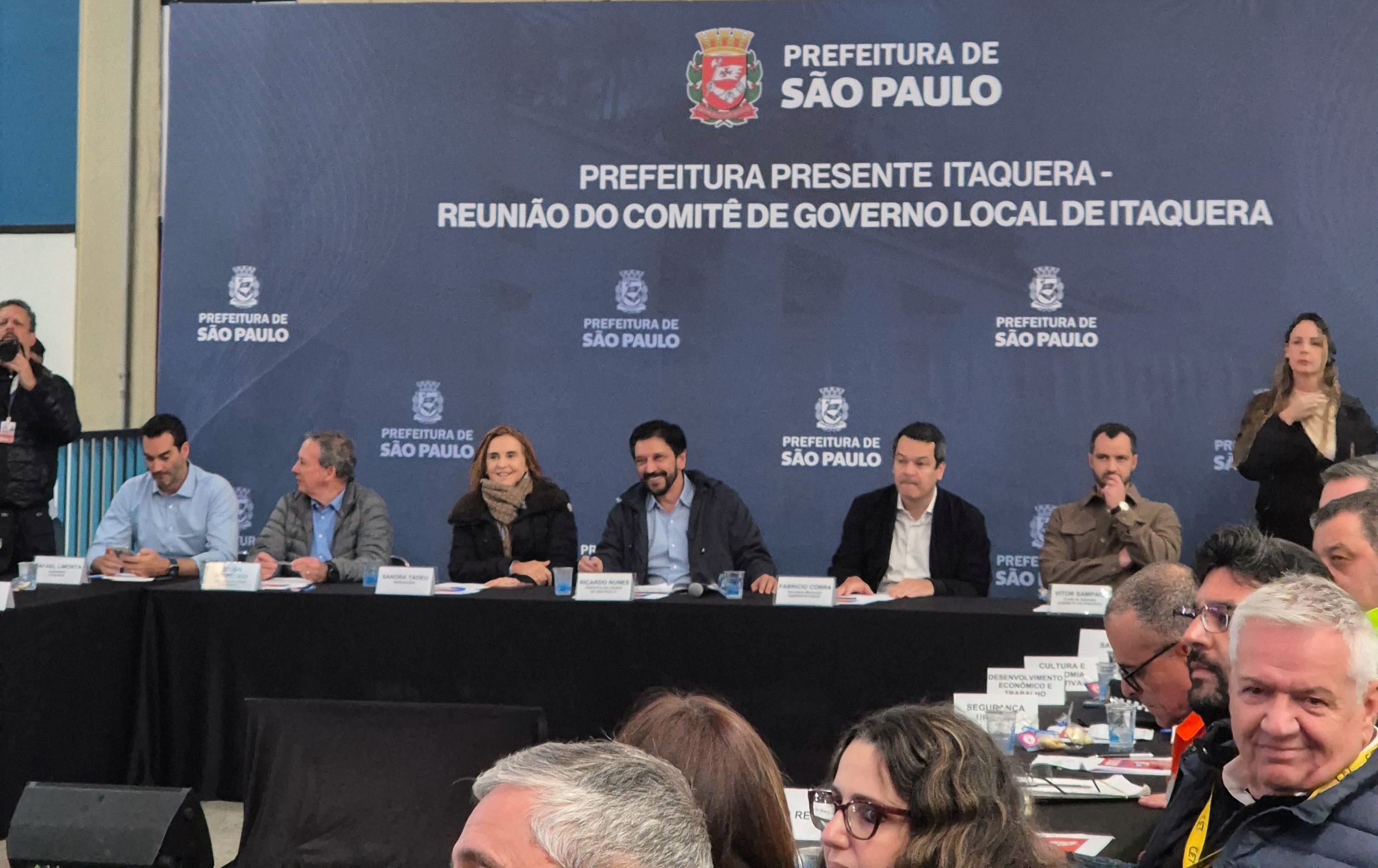 A imagem mostra uma reunião institucional da Prefeitura de São Paulo em Itaquera. Ao centro, há uma longa mesa com várias autoridades sentadas, algumas com microfones à frente. Ao fundo, um painel cinza exibe o logotipo da Prefeitura de São Paulo e o texto "PREFEITURA PRESENTE ITAQUERA - REUNIÃO DO COMITÊ DE GOVERNO LOCAL DE ITAQUERA".  À frente da mesa principal, há cadeiras ocupadas por diversos participantes que acompanham a reunião. Em alguns pontos da mesa, são visíveis garrafas de água e copos. No canto superior direito da imagem, uma mulher loira está de pé, e no canto superior esquerdo, há uma pessoa segurando um microfone.  Em frente a cada participante da mesa principal, há pequenos painéis com o logotipo da "PREFEITURA DE SÃO PAULO". A iluminação do ambiente parece ser boa, e o foco da câmera está nos participantes da mesa principal.
