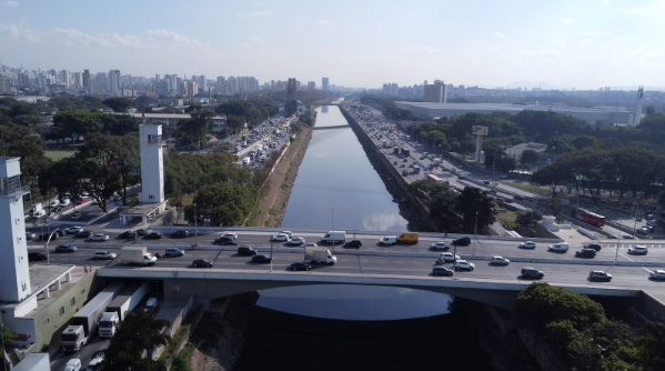 A imagem aérea mostra uma paisagem urbana com um rio ou canal central, ladeado por vias expressas com intenso tráfego de veículos. À esquerda, duas torres brancas se destacam. Ao fundo, arranha-céus e edifícios compõem a linha do horizonte de uma grande cidade. A ponte em primeiro plano está repleta de carros.