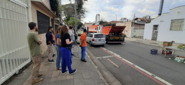 Uma cena de rua mostra um grupo de seis pessoas, a maioria de costas, observando um acidente de trânsito em um bairro urbano. A calçada, à esquerda da imagem, está pavimentada com tijolos de concreto.  Um caminhão de lixo laranja, com a caçamba elevada, está parado no meio da rua. Na frente dele, um carro prata está amassado, indicando a colisão. Próximo ao meio-fio, um homem de camisa laranja e calça jeans está conversando com alguém no interior do carro. Ao lado dele, três pessoas de camisa escura observam a situação. Mais perto da câmera, dois homens de camisa clara, um de barba e um de cabelo escuro, estão de perfil.  Na calçada, mais à frente, há caixas, possivelmente com garrafas e frutas, espalhadas. A rua tem uma faixa vermelha pintada perto do meio-fio e árvores com folhagem verde. No fundo, é possível ver construções e edifícios altos.