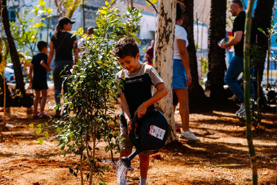 Um menino, de pele clara e cabelo castanho escuro, está de pé no centro da foto, em frente a uma pequena árvore recém-plantada. Ele se inclina para a frente e segura com as duas mãos um regador grande, preto e redondo, derramando água na base da árvore. A cena ocorre ao ar livre, em um espaço de terra batida, com várias outras pessoas desfocadas em segundo plano, incluindo adultos e crianças que observam ou participam do plantio. O sol está brilhando, criando um ambiente claro e iluminado.