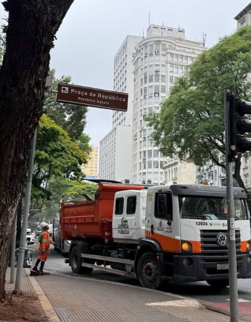 Uma foto de rua em um dia nublado. No canto superior esquerdo, há uma placa de trânsito marrom indicando "Praça da República" e "Republic Square" em letras brancas. À direita da placa e no centro da imagem, está um caminhão branco e laranja de porte médio com a inscrição "LIMPEZA PÚBLICA" na lateral, estacionado na rua. Um funcionário da limpeza pública, vestindo uniforme de segurança laranja e colete refletivo, está em pé na calçada perto da parte traseira do caminhão, olhando para a frente. O lado esquerdo da imagem é ladeado por árvores, uma das quais tem um tronco grosso e escuro em primeiro plano. Ao fundo, vê-se a paisagem urbana com edifícios altos, brancos e claros, característicos do centro de uma cidade, sob um céu cinzento.