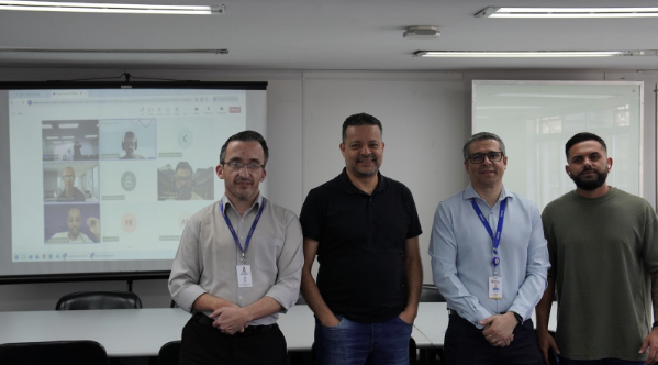 Quatro homens adultos estão lado a lado em uma sala de reunião. Três deles usam camisas sociais ou polo, e um usa uma camiseta casual. No fundo, à esquerda, uma tela de projeção exibe uma videochamada ativa com múltiplos participantes em miniatura.