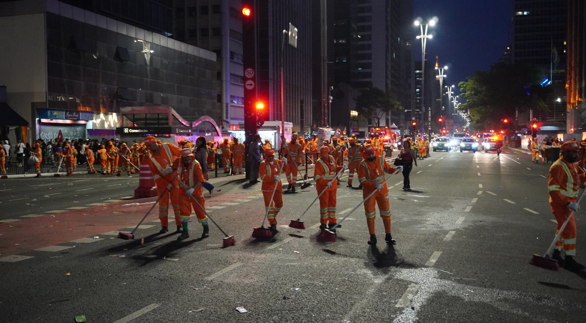 A fotografia noturna mostra um grupo de garis varrendo uma rua larga e asfaltada, que parece ser uma avenida movimentada. A cena é dominada pela presença de muitos trabalhadores, todos vestidos com uniformes laranja vibrante e coletes refletivos, segurando vassouras longas. Eles estão espalhados pela pista, que está molhada em algumas partes, refletindo as luzes da cidade.  Ao fundo, há vários prédios altos, característicos de uma área urbana central, com suas janelas iluminadas. Semáforos vermelhos podem ser vistos, indicando que o tráfego está parado ou que é tarde da noite. À direita da imagem, alguns carros estão parados na rua, e suas luzes traseiras vermelhas e faróis brancos brilham. Há postes de luz com luminárias acesas, iluminando a cena. A iluminação artificial e os uniformes dos garis se destacam no ambiente escuro da noite. O chão está com alguns resíduos, indicando que a limpeza está em andamento.