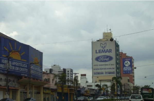 Uma imagem de rua em uma cidade, com prédios de vários andares e placas de publicidade. Na parte esquerda, há um grande cartaz com um fundo azul e um sol amarelo com raios, com os dizeres "IGREJA APOSTÓLICA RENASCER EM CRISTO". No centro, um prédio branco e alto tem um painel azul com o nome "LEMAR" em branco e o logo da Ford. Abaixo, há a frase "ESTE TERRITÓRIO É SEU". À direita, há outro cartaz vertical, azul e branco. O céu está nublado, e o trânsito na rua, com carros e ônibus, parece moderado.