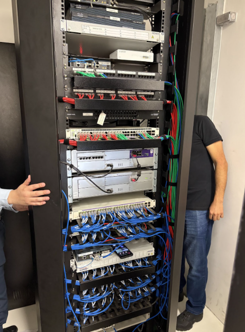 A imagem mostra um rack de servidores e equipamentos de rede preto, preenchido com diversos dispositivos, como switches e patch panels. Há uma grande quantidade de cabos de rede, predominantemente azuis, que estão conectados aos equipamentos de forma organizada. É possível ver alguns cabos vermelhos e, à direita, um feixe de cabos de cores variadas (verde, vermelho e azul) descendo ao lado do rack. À direita do rack, é visível parte de um homem vestindo uma camiseta preta e calça jeans, com o rosto fora do enquadramento, e à esquerda, uma mão estendida de outra pessoa.