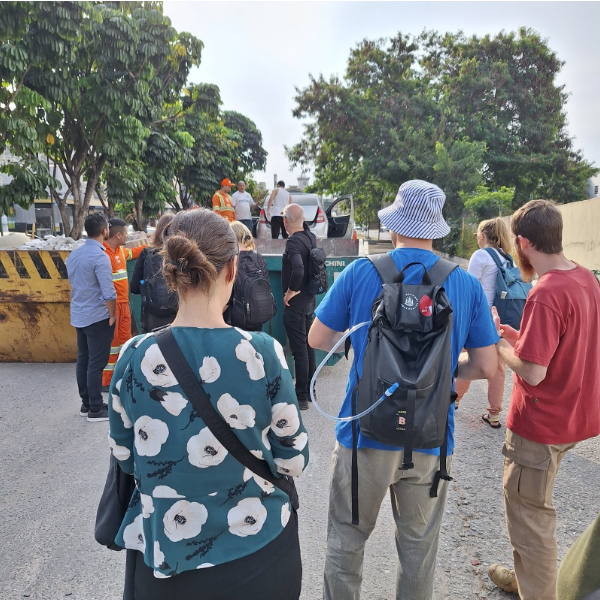 A imagem mostra um grupo de pessoas de costas, observando uma cena. Elas estão em um pátio ou rua. Em primeiro plano, uma mulher com uma blusa de estampa floral, e ao seu lado um homem usando um chapéu listrado, com uma mochila preta e um tubo de hidratação.  Mais ao fundo, um grupo de pessoas, incluindo alguns trabalhadores com coletes de segurança laranjas, está em volta de uma caçamba de lixo metálica, de cor verde. O grupo parece estar conversando e alguns estão apontando. Uma parte de um carro branco também é visível próximo à caçamba.  A cena é cercada por árvores, e há um muro em segundo plano. No canto inferior esquerdo, uma parte de uma estrutura de metal amarela está visível.