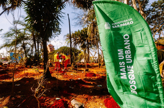 Uma bandeira verde, em primeiro plano à direita, anuncia o projeto "Mais um Bosque Urbano", da Prefeitura de São Paulo. Ao fundo, em uma área de terra avermelhada, três trabalhadores com uniformes laranjas e coletes amarelos estão plantando mudas de árvores em meio a vegetação rasteira e algumas palmeiras.