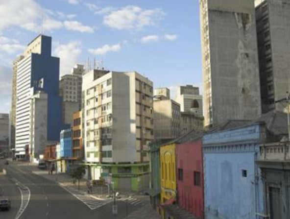 Uma rua de cidade movimentada, com prédios de várias alturas e estilos. À esquerda, um arranha-céu com a fachada azul e branca se destaca entre os demais. No centro, há um prédio residencial de cinco andares, e na parte inferior, pequenos edifícios comerciais coloridos, pintados de azul, laranja, verde, amarelo e rosa. A rua está vazia, com poucos carros em movimento. O céu está parcialmente nublado, com nuvens brancas e o sol.