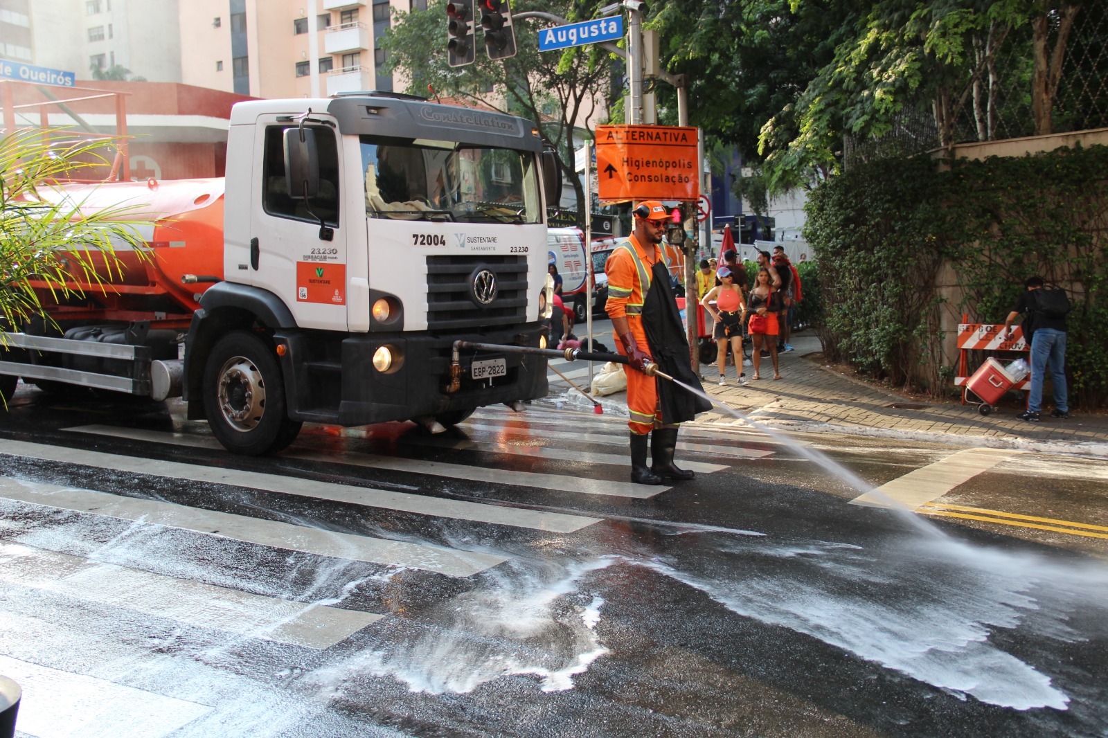 Uma imagem colorida exibe uma cena de limpeza urbana em São Paulo, Brasil. No primeiro plano, um caminhão-pipa branco com detalhes laranja está parado sobre uma faixa de pedestres branca, inclinando-se ligeiramente para a direita da foto. O caminhão tem o número "72004" visível na lateral e um adesivo laranja e branco.  À direita do caminhão, um funcionário da limpeza pública, vestindo um uniforme laranja, colete preto, luvas e botas pretas, segura uma mangueira e direciona um jato d'água potente sobre o asfalto, que aparece molhado e espumoso. O homem tem um rádio preso ao colete.  Ao fundo, é possível ver parte de um prédio, árvores, um semáforo, e uma placa de trânsito azul com a palavra "Augusta" em destaque. À direita da imagem, outras pessoas observam a cena, algumas vestindo roupas coloridas, e ao fundo, um homem de calça jeans e camisa azul escura está agachado. A luz do sol incide sobre a cena, criando um contraste de luz e sombra.