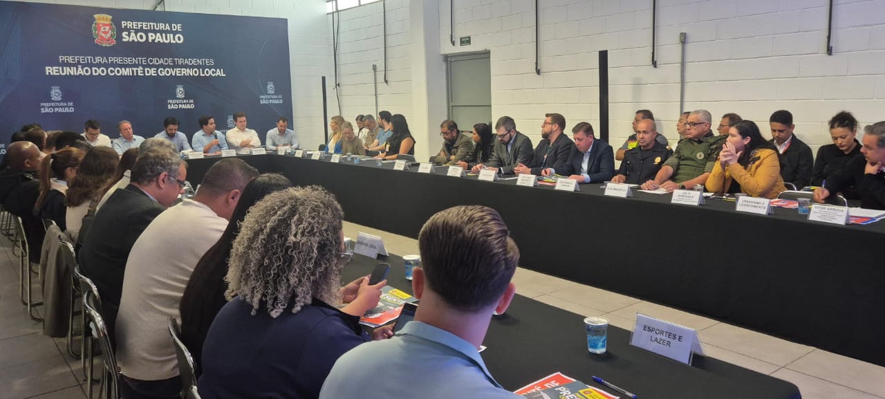 A foto registra uma reunião de trabalho em ambiente fechado, com dezenas de pessoas sentadas em torno de uma longa mesa de toalha preta. Ao fundo, um grande painel institucional da Prefeitura de São Paulo, Subprefeitura Cidade Tiradentes, anuncia o evento como a "REUNIÃO DO COMITÊ DE GOVERNO LOCAL". Os participantes, incluindo autoridades de terno e uniformes militares ou da Guarda, estão distribuídos em ambos os lados da mesa, focando a atenção nos que estão sentados na cabeceira. No primeiro plano, à esquerda, é possível ver as costas e a nuca de dois participantes, sendo um deles com cabelos loiros e cacheados, olhando para o celular. O ambiente é formal e bem iluminado.