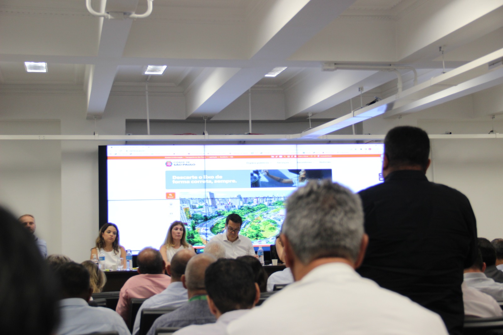 A imagem mostra um evento ou reunião em um ambiente corporativo, com um painel de palestrantes à frente de uma grande tela exibindo um site da Prefeitura de São Paulo. O público está atento, com um homem de preto em pé, possivelmente fazendo uma pergunta ou comentário. Os palestrantes incluem duas mulheres e um homem, que parecem estar conduzindo a apresentação. O local tem um design moderno, com iluminação clara e estrutura bem organizada, sugerindo um evento formal de discussão ou planejamento.