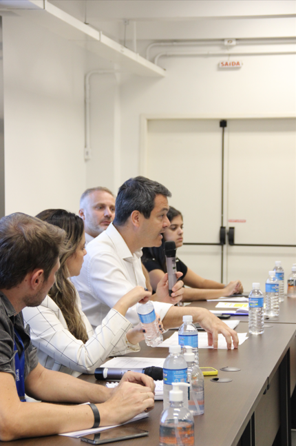 A imagem mostra um grupo de pessoas participando de uma reunião em uma sala bem iluminada. Um homem de camisa branca segura um microfone e fala enquanto gesticula com a mão. Outros participantes, incluindo uma mulher de cabelo longo e um homem de camisa preta, prestam atenção à discussão. Sobre a mesa, há garrafas de água, papéis, canetas e celulares. Ao fundo, uma porta branca com uma placa vermelha indicando "SAÍDA" pode ser vista. A cena sugere um ambiente corporativo ou institucional, com foco na comunicação e no debate de ideias.