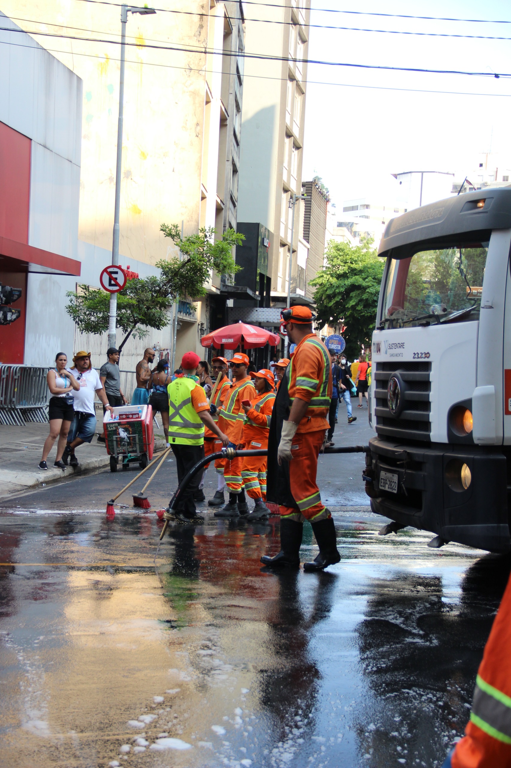 Uma imagem vertical colorida mostra uma rua urbana em São Paulo, Brasil, após uma ação de limpeza. À direita da foto, parte de um caminhão de lixo branco é visível. O asfalto no primeiro plano está molhado e reflete a luz, com algumas manchas de espuma branca.  No centro da imagem, um grupo de funcionários da limpeza pública, vestidos com uniformes laranja vibrante e coletes refletivos, está trabalhando. Um deles, de costas para o observador e vestindo um colete verde-limão, utiliza uma vassoura para mover a água na rua. Outro funcionário, mais próximo do caminhão, veste um boné laranja e luvas escuras, observando a cena.  À esquerda, um grupo de pessoas, algumas com roupas esportivas, está parado perto de uma parede vermelha e branca, observando a atividade. Ao fundo, prédios altos e a vegetação das árvores se destacam sob o céu claro. Há também uma placa de proibido estacionar (símbolo E cortado) visível à esquerda. A cena transmite uma sensação de atividade e trabalho em equipe para manter a cidade limpa.