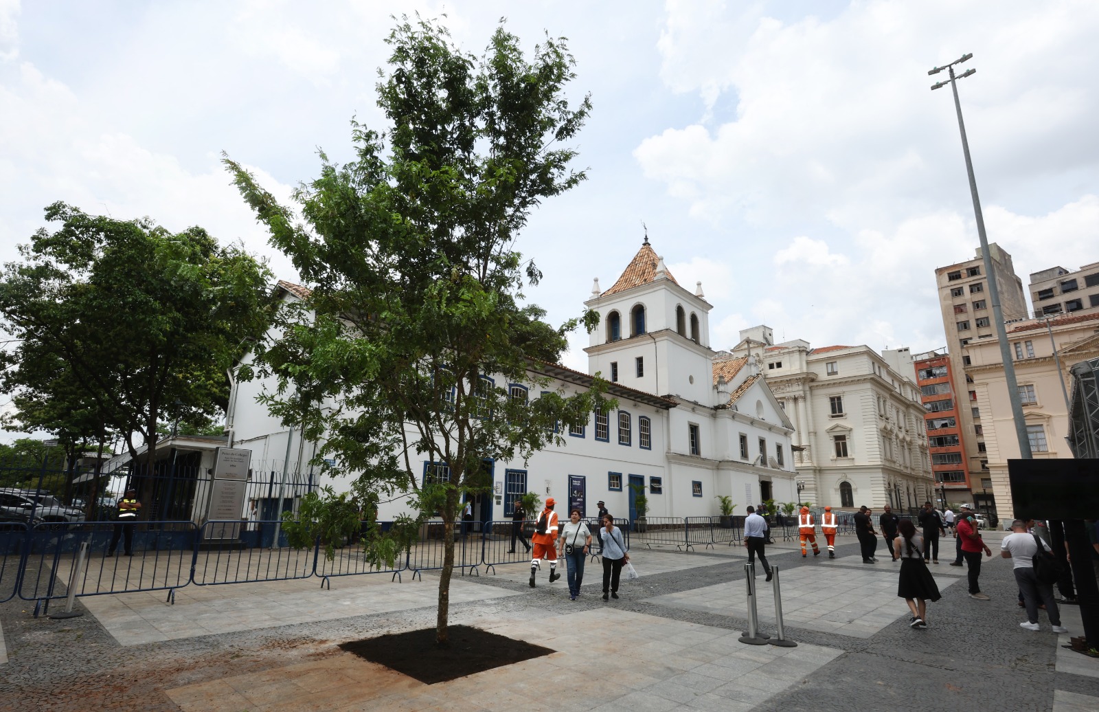 A imagem, tirada em um dia claro, mostra uma praça ou largo com piso de pedra em tom cinza claro. Em primeiro plano, no centro, há uma árvore jovem com tronco fino e copa verde, plantada em um quadrado de terra.  Ao fundo, domina a cena um edifício histórico de cor branca, com telhado de telhas e detalhes em azul nas janelas. O prédio tem uma torre central com telhado em formato cônico e uma cruz no topo. À esquerda do prédio, há árvores grandes e densas e uma área cercada por grades escuras.  Na parte inferior da foto, várias pessoas caminham ou estão paradas na praça, incluindo algumas vestindo uniformes de segurança ou serviço em tons de laranja e preto. À direita e mais ao fundo, observam-se prédios modernos de múltiplos andares. Um poste de iluminação alto se eleva no canto superior direito. O céu está parcialmente nublado.