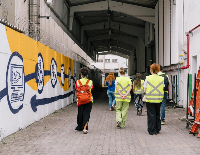 Foto de um grupo de pessoas caminhando em fila indiana em um corredor externo de um prédio industrial. As pessoas estão de costas para a câmera, usando coletes amarelos com detalhes refletivos. A primeira pessoa da fila usa uma mochila laranja. À esquerda, há um muro com grafites de ícones brancos em um fundo amarelo e azul, incluindo um computador, um tênis e uma bicicleta. À direita, há uma escada metálica laranja encostada na parede. O corredor é longo e leva a uma área com mais prédios ao fundo. O dia parece nublado, com luz difusa iluminando a cena.