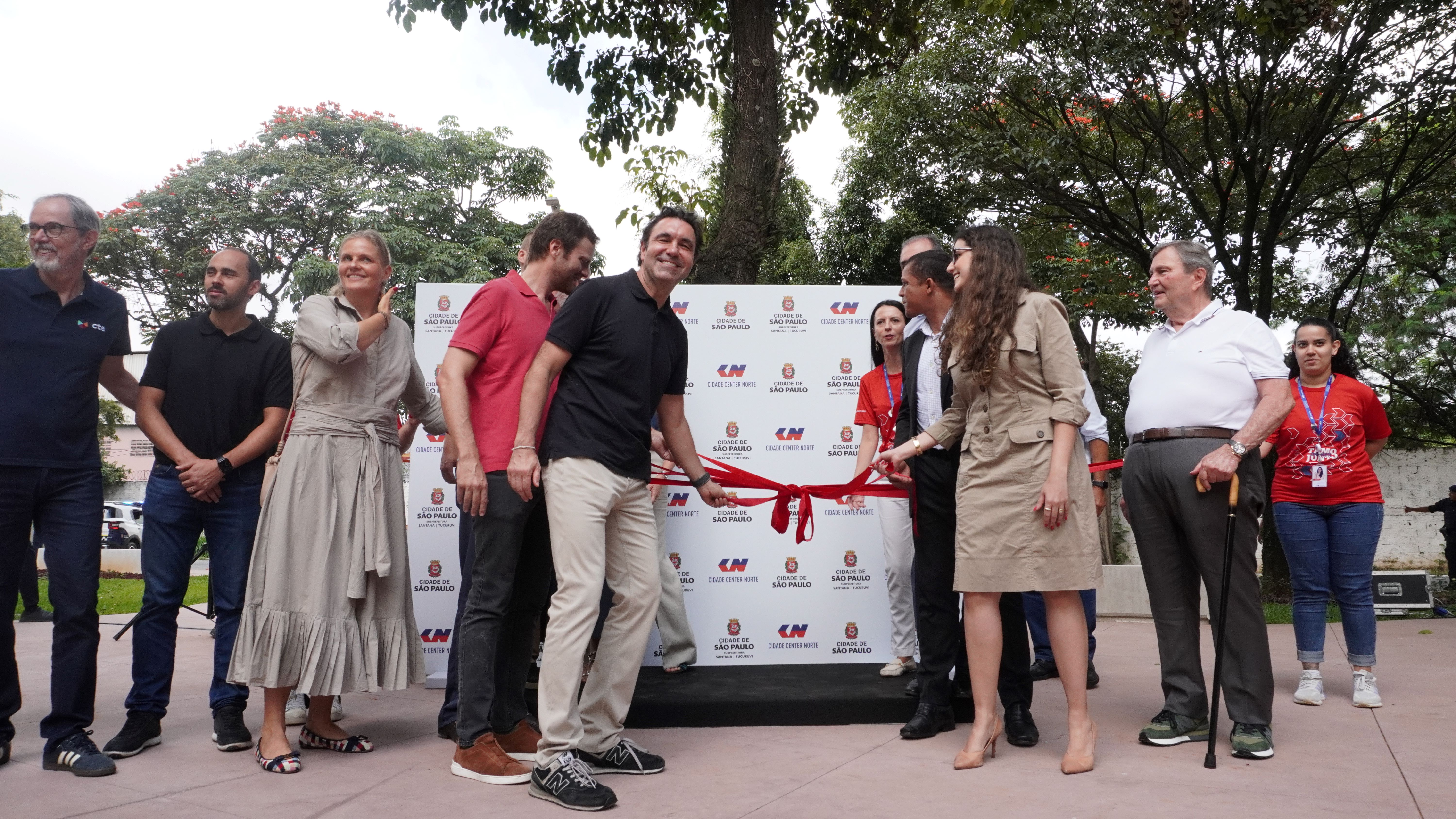 Grupo de pessoas participando de uma cerimônia de inauguração ao ar livre, realizando o tradicional corte da fita vermelha. No centro da imagem, algumas pessoas seguram a fita e sorriem para a câmera, enquanto outras ao redor observam o momento. Ao fundo, há um painel institucional com logotipos da Prefeitura de São Paulo e de empresas envolvidas no evento. A cena acontece em um ambiente arborizado, com árvores e gramado, sugerindo a inauguração de um espaço público, como uma praça ou parque.