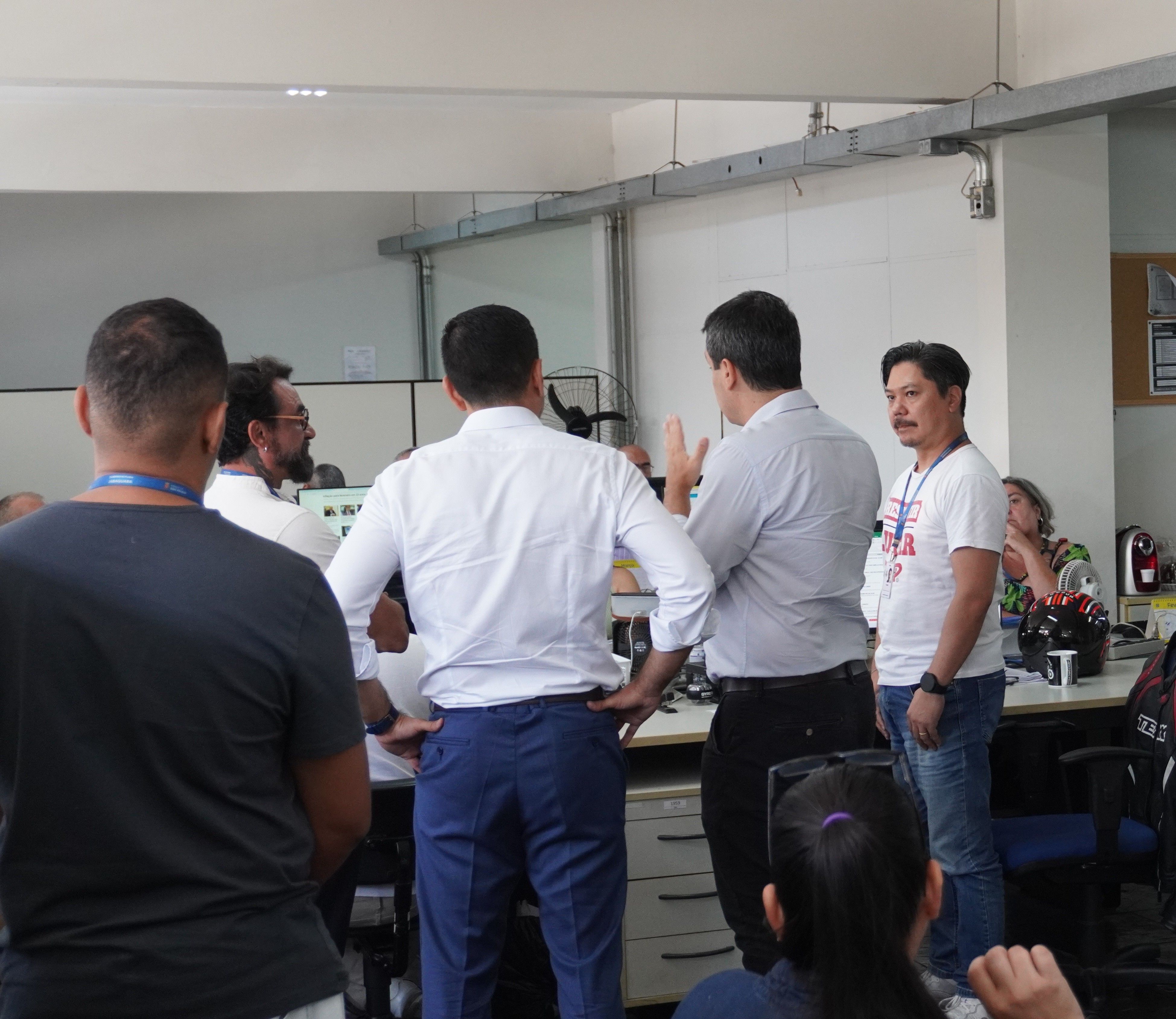 A imagem mostra um grupo de pessoas reunidas em um ambiente de trabalho, possivelmente um laboratório ou escritório técnico. Algumas pessoas estão de costas para a câmera, enquanto outras estão de perfil ou de frente, participando de uma conversa. Um homem de camisa cinza e calça escura parece estar explicando algo, gesticulando com as mãos, enquanto os demais escutam atentamente. O ambiente conta com mesas, equipamentos e documentos, sugerindo um espaço de trabalho colaborativo. Algumas pessoas estão sentadas, enquanto outras estão de pé, reforçando a dinâmica interativa da cena.
