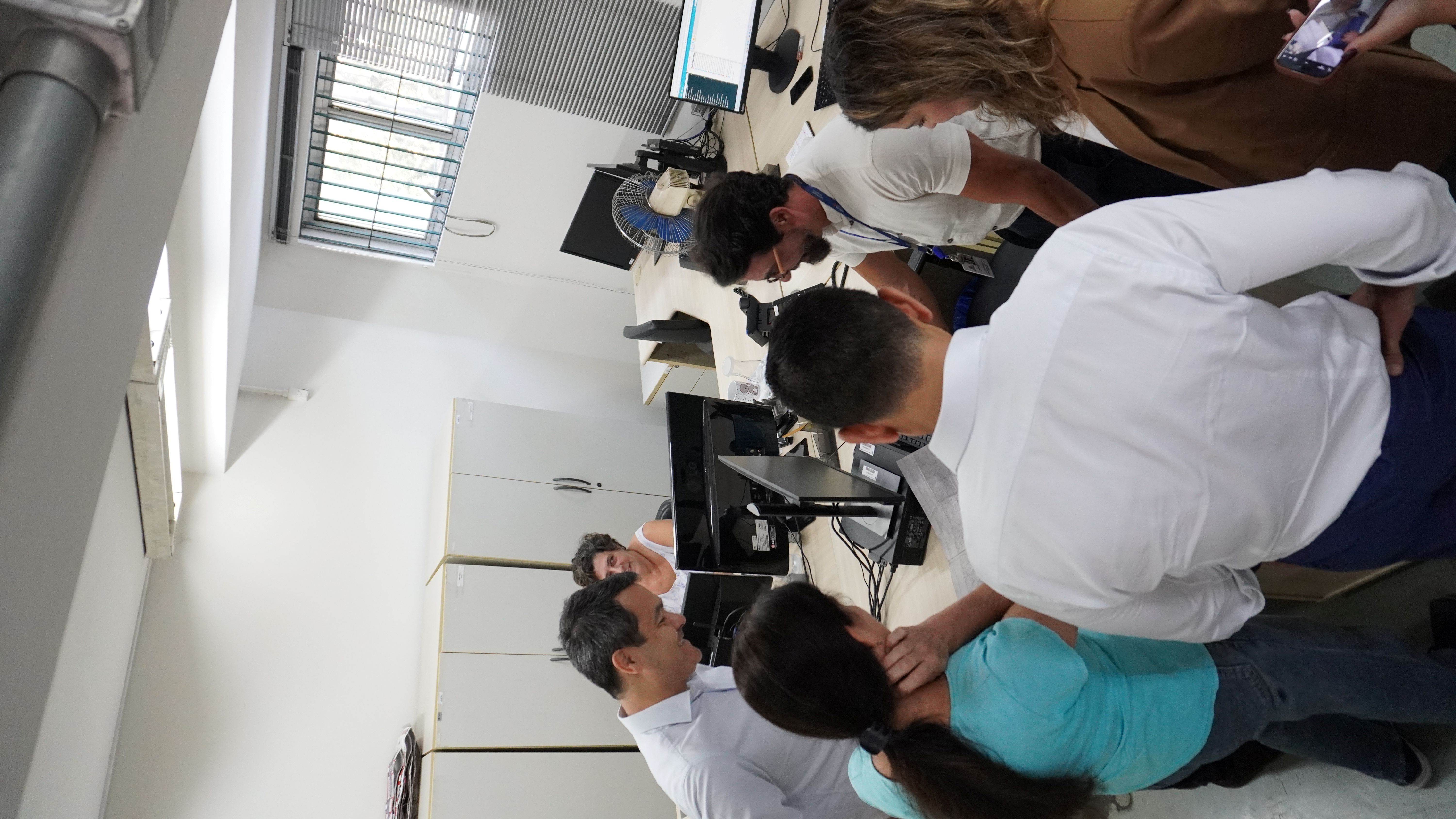  VER Imagem de um grupo de pessoas trabalhando em equipe. Elas estão em um escritório, algumas sentadas e outras em pé, conversando e olhando para computadores. As pessoas parecem engajadas e colaborativas. O ambiente é iluminado e há móveis de escritório visíveis.