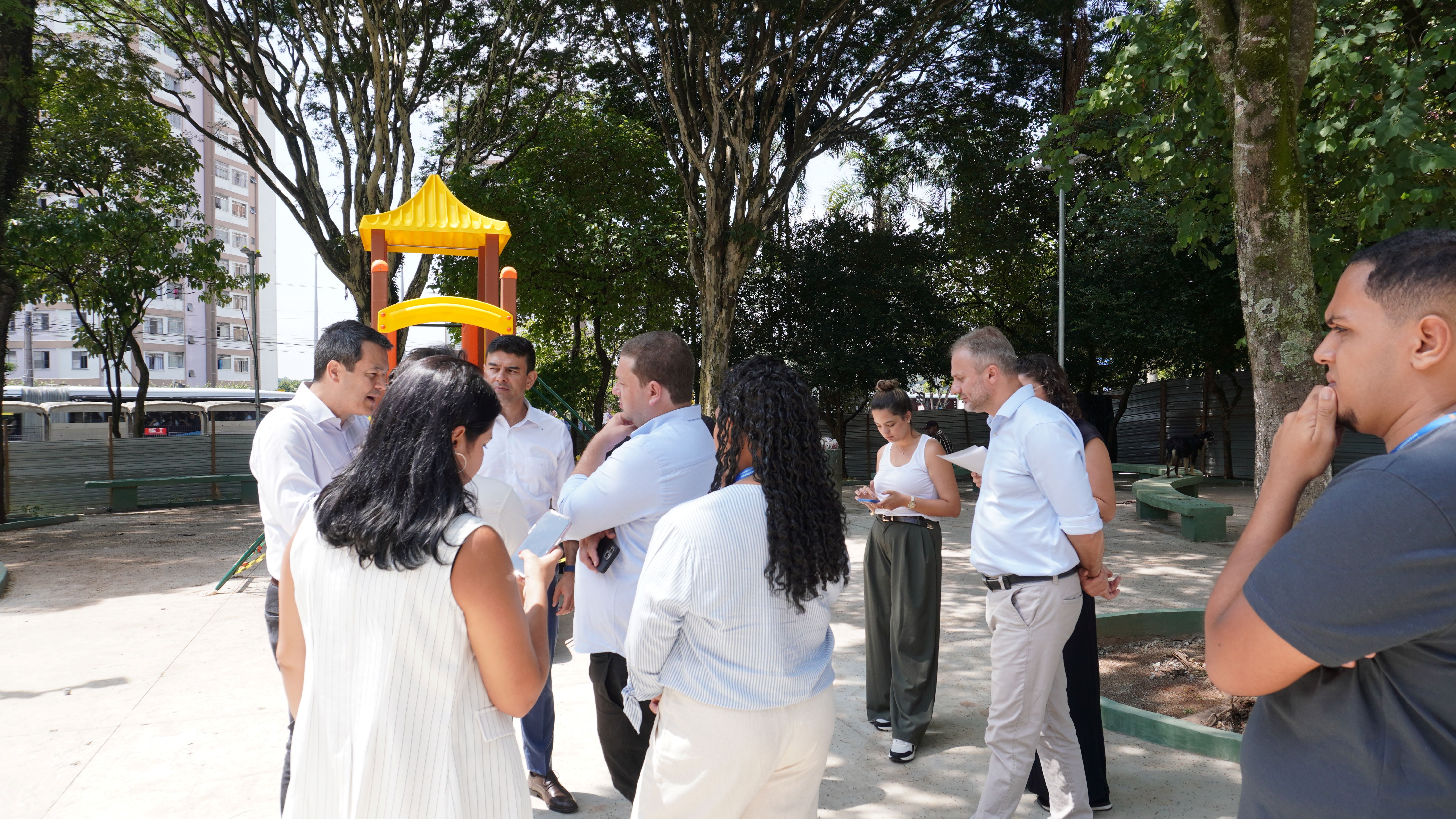 Foto de um grupo de pessoas reunidas ao ar livre, em um parque com árvores e um playground infantil ao fundo. As pessoas estão em pé, conversando e interagindo. A maioria está vestida com roupas casuais, em tons claros. Ao fundo, árvores altas e verdes criam uma sombra fresca, e um playground colorido com escorregador amarelo se destaca. A luz do dia ilumina a cena, criando um ambiente aberto e convidativo.