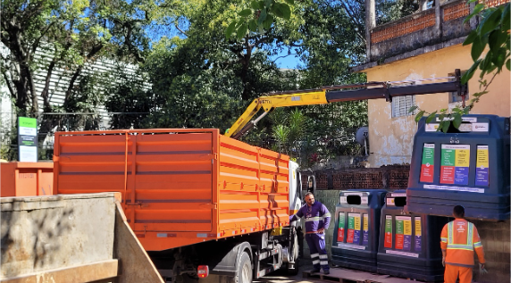 Na imagem, um caminhão laranja equipado com um braço hidráulico realiza a coleta de resíduos em um ecoponto. O braço mecânico está em operação, aparentemente esvaziando um dos contentores verdes de coleta seletiva, que possuem adesivos coloridos indicando os tipos de materiais recicláveis. Dois trabalhadores uniformizados estão presentes: um manuseando o equipamento e outro próximo aos contentores, possivelmente auxiliando na operação. Ao fundo, há uma construção amarela e vegetação densa, sugerindo um ambiente urbano arborizado. O dia está ensolarado, com céu azul e sombras projetadas pelas árvores.