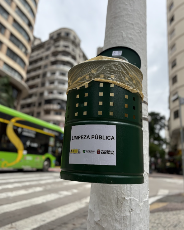 A imagem mostra, em primeiro plano, uma lixeira pública cilíndrica de metal na cor verde escura, fixada em um poste branco de textura rugosa. A lixeira possui pequenos furos quadrados em sua parte superior, revelando um saco plástico amarelo no interior. No centro do objeto, há um adesivo branco com a frase "LIMPEZA PÚBLICA" e logotipos da Prefeitura de São Paulo e de empresas de saneamento. O cenário ao fundo, levemente desfocado, apresenta uma rua com faixa de pedestres, um ônibus urbano verde em movimento à esquerda e grandes edifícios residenciais de arquitetura antiga sob um céu nublado.