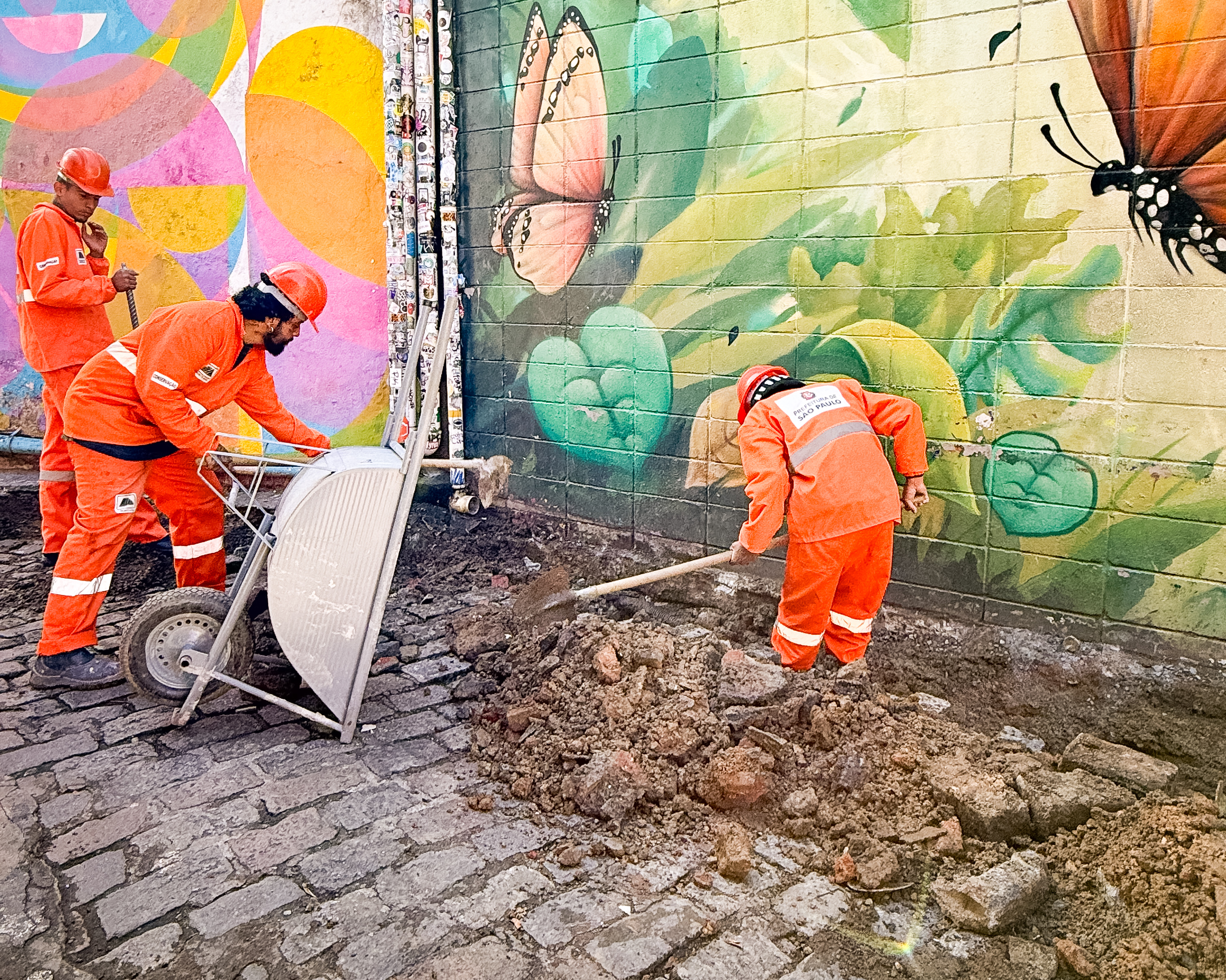 Imagem mostra trabalhadores da Prefeitura de São Paulo executando obras de requalificação no Beco do Batman. As equipes utilizam ferramentas manuais para escavação e manutenção da via de paralelepípedos ao lado de murais coloridos com desenhos de borboletas e formas geométricas. Os profissionais usam uniformes laranja e equipamentos de segurança durante a realização das melhorias no espaço urbano.