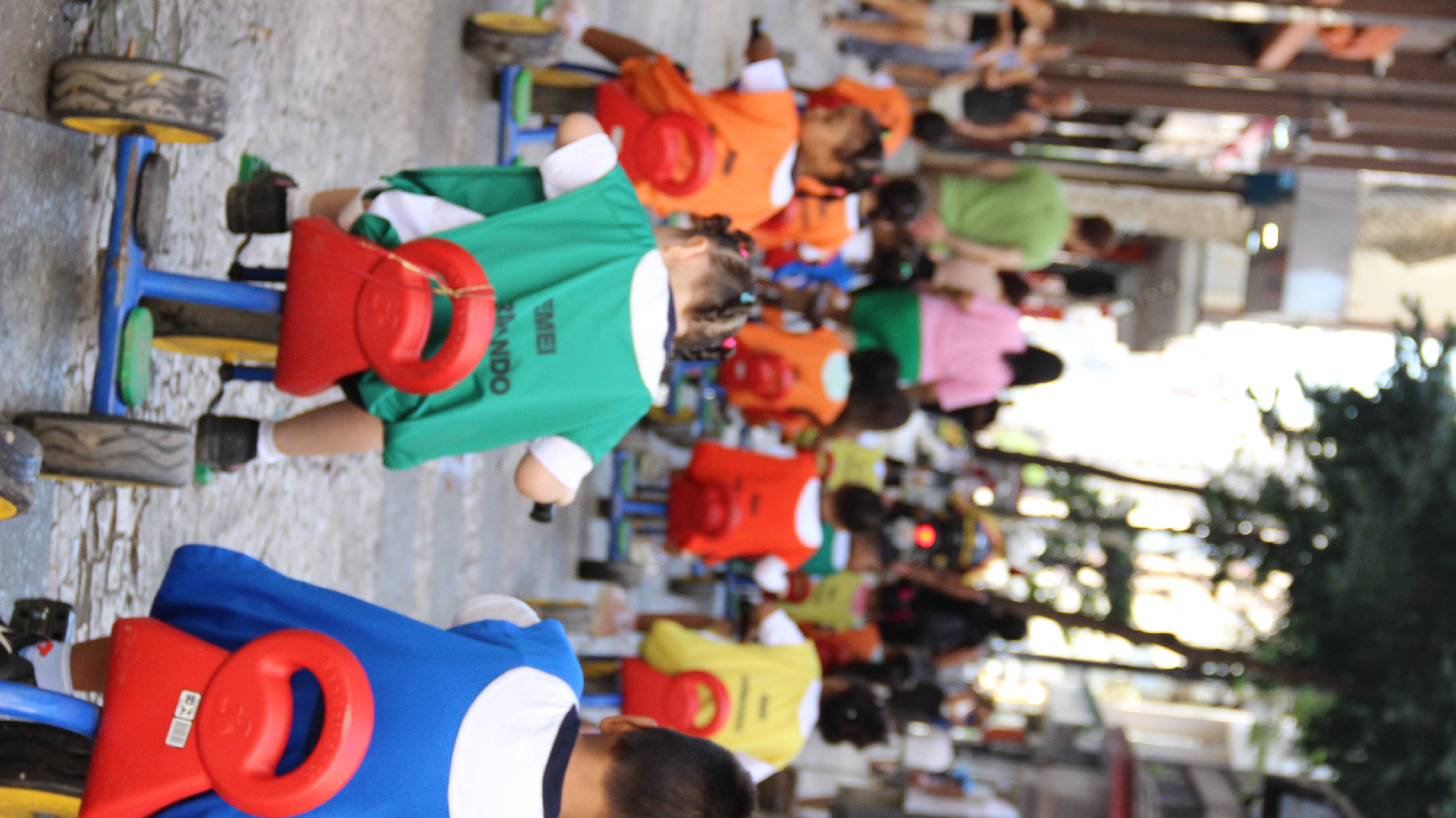 A imagem mostra um grupo de crianças pequenas andando de triciclo em uma rua de calçamento. Elas vestem coletes coloridos, possivelmente identificando a instituição escolar à qual pertencem, como indicado pelo texto "EMEI" visível nas costas de algumas delas. A cena acontece em um ambiente urbano movimentado, com adultos caminhando ao fundo e prédios comerciais e residenciais ao redor. A iluminação natural e a presença de sombras sugerem que a atividade ocorre durante o dia.