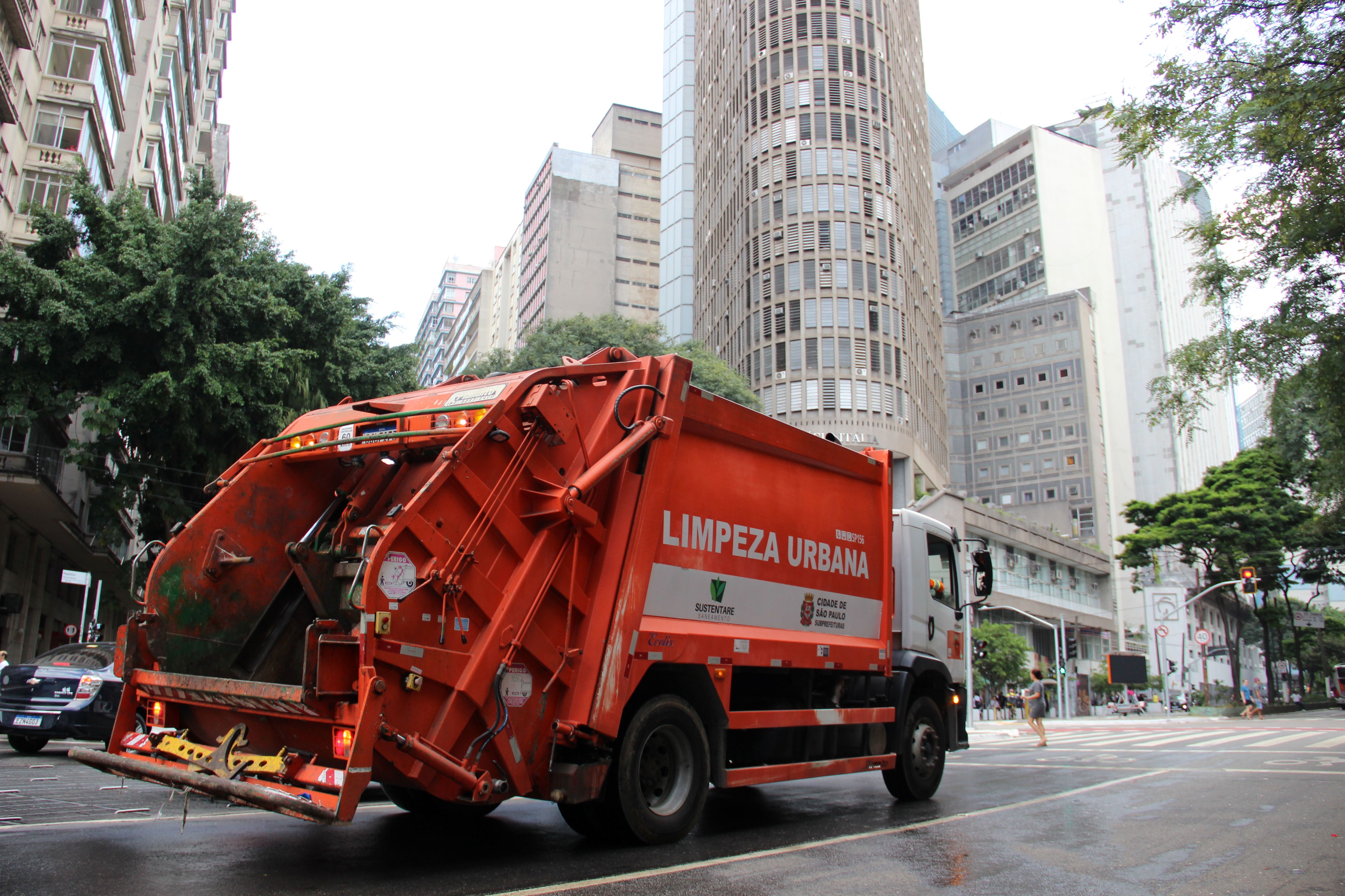 Um caminhão de coleta de lixo de cor laranja, identificado com a inscrição "LIMPEZA URBANA", circula por uma rua movimentada em uma área central da cidade. O veículo pertence ao serviço de limpeza da Prefeitura de São Paulo, conforme indicado nas laterais. No fundo, prédios altos e modernos, árvores e alguns pedestres atravessando a rua compõem a paisagem urbana. A via apresenta sinais de umidade, sugerindo que pode ter chovido recentemente ou passado por um processo de limpeza.