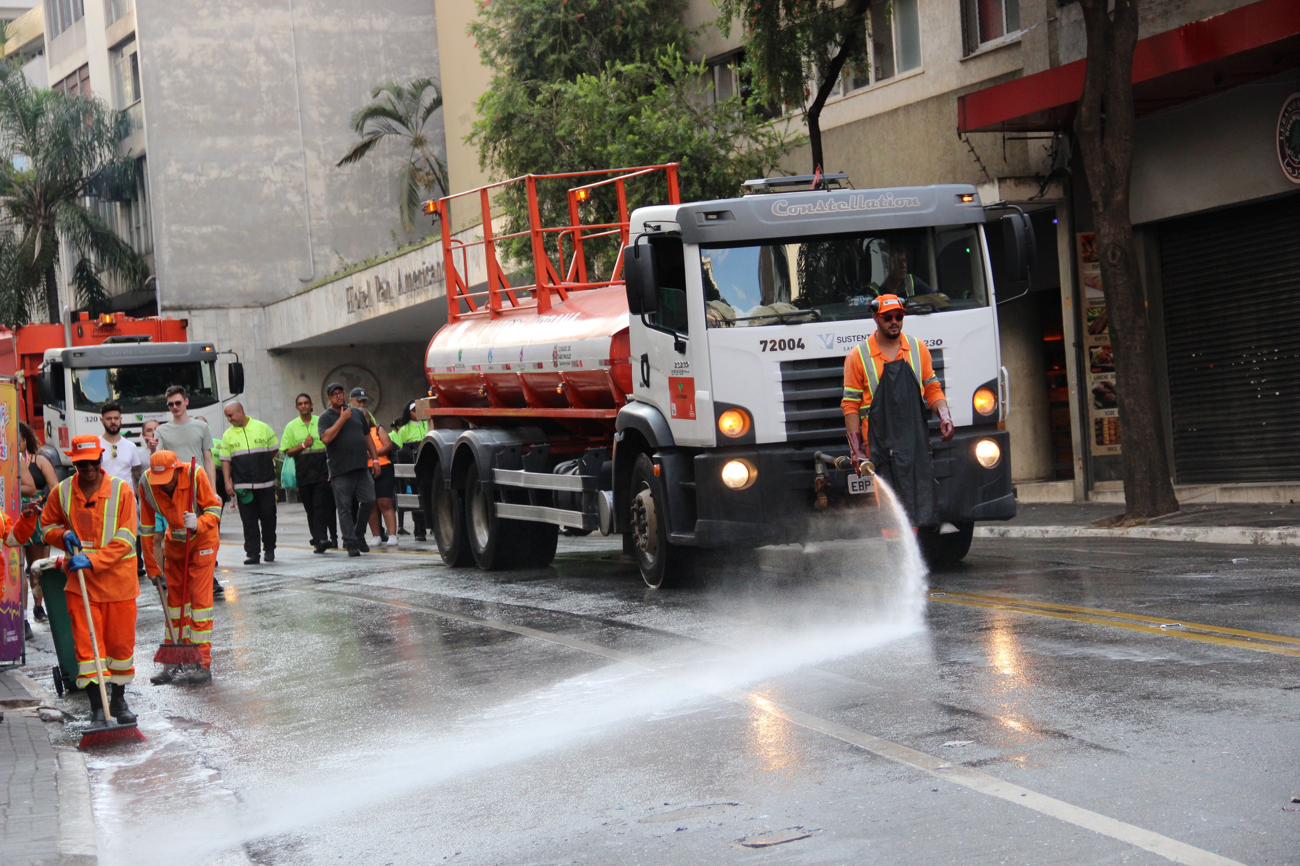 Trabalhadores com uniformes laranja de alta visibilidade realizam a limpeza de uma rua urbana durante o dia. Um caminhão-pipa está estacionado na via, e um dos trabalhadores, usando um avental de borracha e luvas, opera uma mangueira de alta pressão para lavar o asfalto. Outros funcionários varrem a água para as laterais da rua. No fundo, há prédios, árvores e pedestres observando a operação, alguns vestindo coletes de segurança. O ambiente urbano e a presença de caminhões indicam uma possível manutenção ou higienização da via pública.