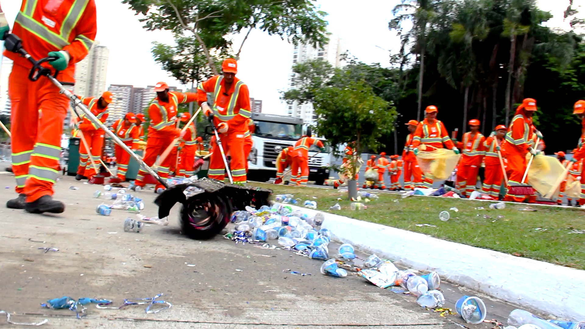 Uma equipe de trabalhadores da limpeza urbana, vestidos com uniformes laranja e faixas refletivas, realiza a varrição e coleta de lixo em uma área pública após um grande evento. No chão, há uma grande quantidade de copos plásticos, garrafas e outros resíduos espalhados. Um dos trabalhadores utiliza uma ferramenta para juntar o lixo, enquanto outros seguram sacos plásticos amarelos para recolher os resíduos. Ao fundo, é possível ver mais trabalhadores atuando, um caminhão de coleta e edifícios altos, indicando um ambiente urbano movimentado.