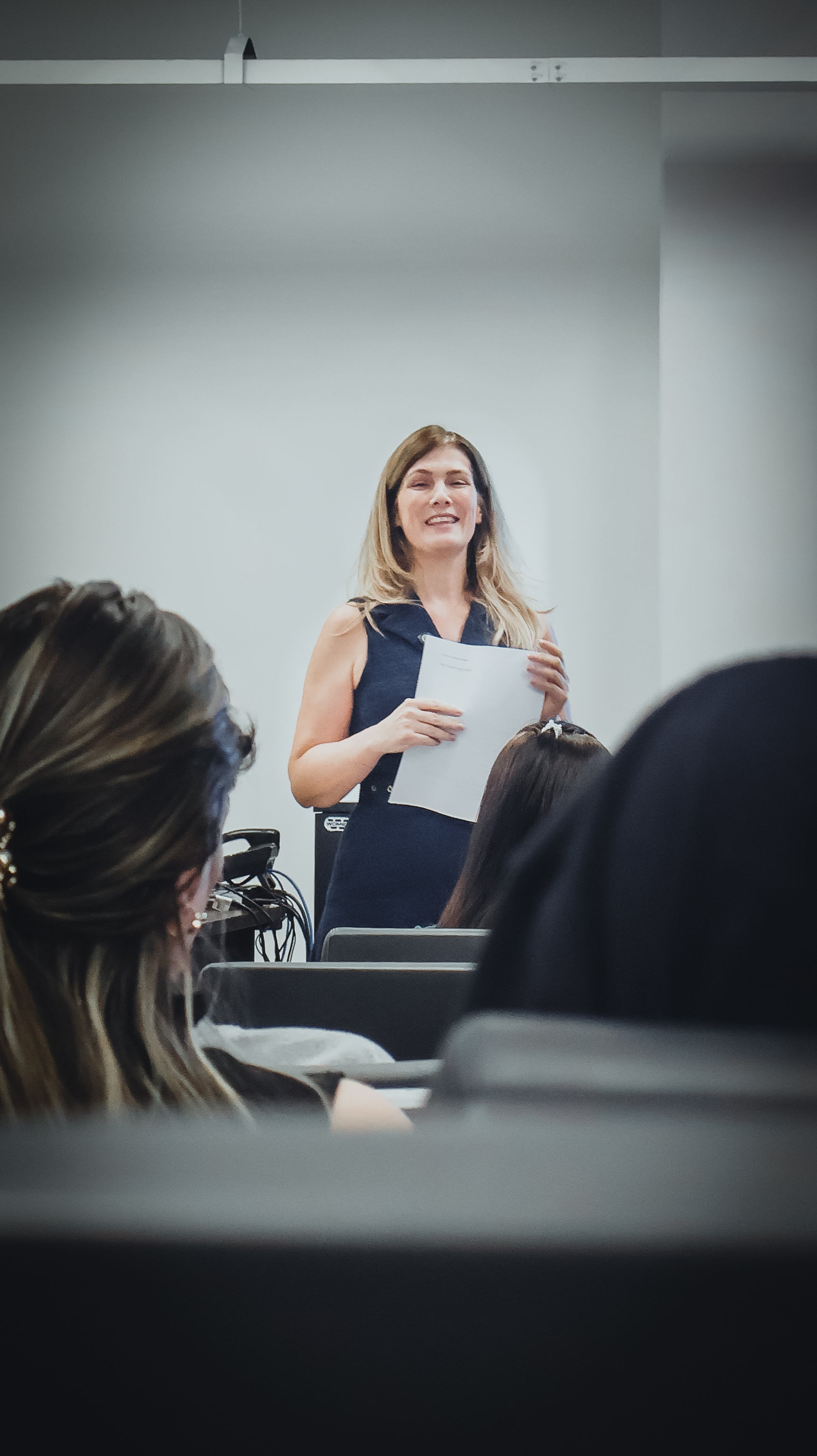 Uma mulher de cabelo loiro, sorridente e vestindo um vestido azul marinho sem mangas, está de pé no centro de um palco de auditório, segurando um maço de documentos. Ela está olhando para a frente, em direção a um público invisível. A foto é tirada do ponto de vista da platéia, com as silhuetas desfocadas e as costas das cabeças de vários membros do público preenchendo o primeiro plano inferior e as laterais, emoldurando a palestrante. O fundo consiste em uma parede branca simples com estruturas de trilho de iluminação visíveis no teto. O foco está nítido na mulher, enquanto o público e o fundo estão desfocados. O evento parece ser uma apresentação, cerimônia de formatura ou entrega de prêmios, com uma atmosfera calorosa e profissional.