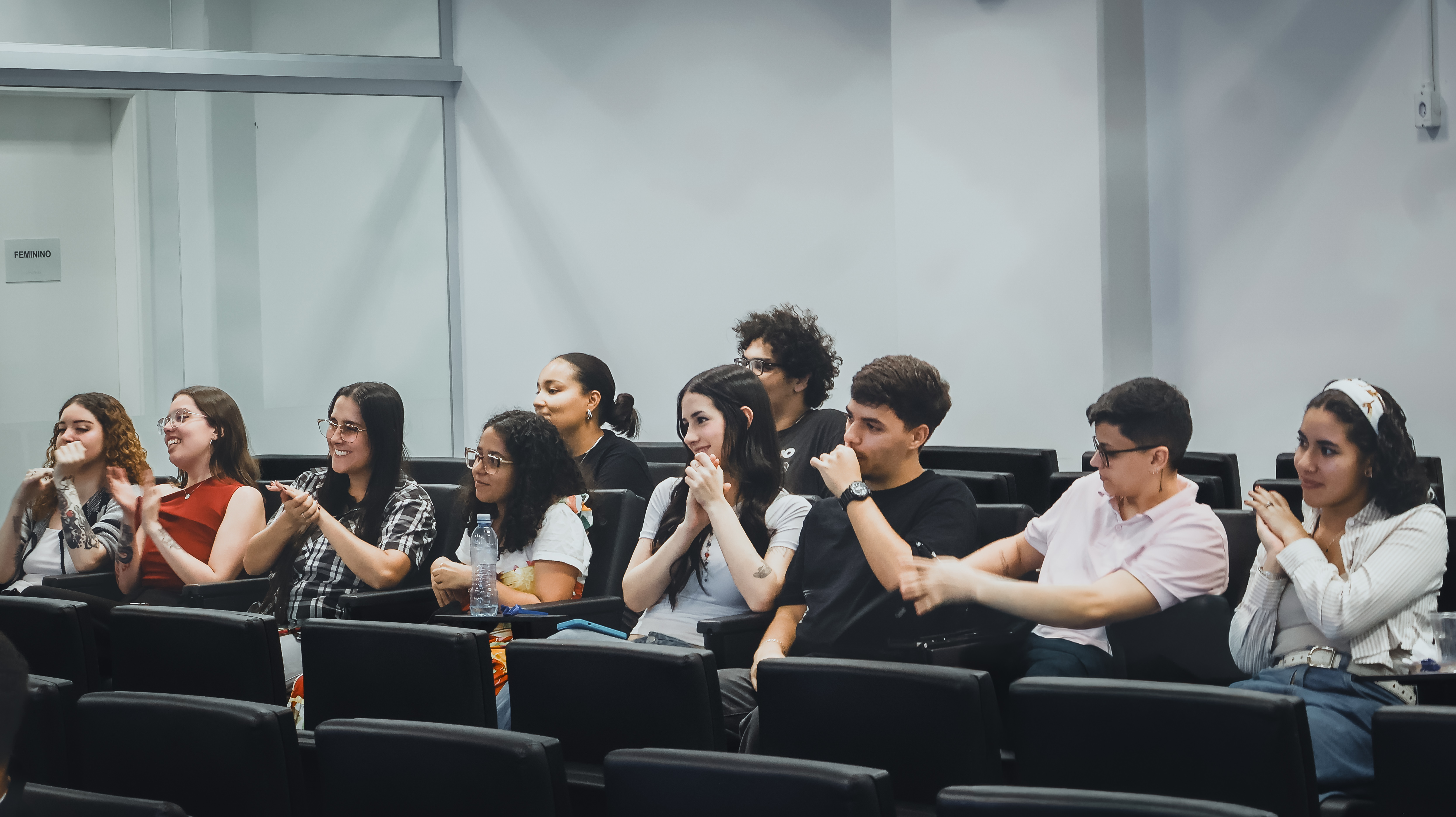 A imagem captura um grupo de nove jovens adultos sentados em poltronas pretas de um auditório, demonstrando um entusiasmo genuíno e foco em algo que acontece fora do enquadramento, à esquerda. A atmosfera é de engajamento coletivo, com a maioria dos presentes sorrindo abertamente e batendo palmas. Na fila da frente, a diversidade de estilos é marcante: vemos jovens de óculos, uma moça com tatuagens visíveis nos braços, outra vestindo uma camisa xadrez e um rapaz de camiseta preta que observa a cena com a mão próxima ao rosto, em sinal de concentração.  Ao fundo, a segunda fileira completa a composição com um rapaz de camisa rosa claro e uma jovem de faixa no cabelo, ambos compartilhando do mesmo clima positivo. O ambiente possui um aspecto institucional ou acadêmico, com paredes em tons neutros e uma divisória de vidro ao fundo onde se lê a placa "Feminino". A iluminação clara e uniforme realça as expressões de interesse e a descontração do grupo, transformando o registro em um momento vibrante de aprendizado ou celebração.